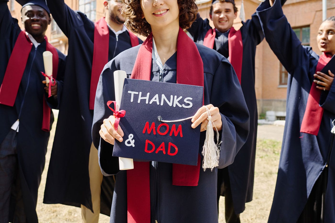 Portrait of graduated student holding hat with text, she thanking her parents together with other students in background